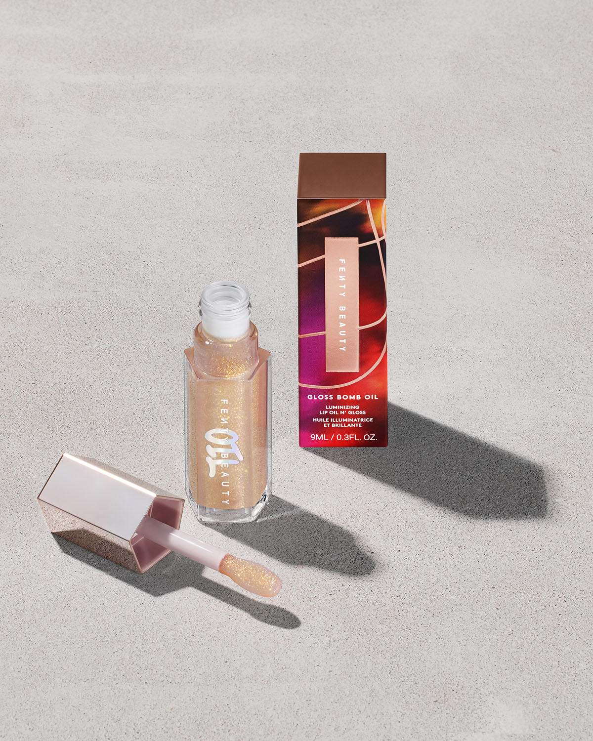 Fenty Beauty Gloss Bomb Oil in shimmering gold with box on a light textured surface